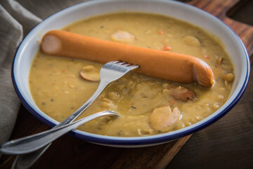 Deftige Deutsche Erbsensuppe als Eintopf mit Bockwurst in Emaille Teller auf Holzbrett und Holz  Hintergrund dunkel