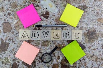Advert or advertising concept with multicolored memo pads, pen and magnifying glass surrounding central alphabet letters on wooden cubes over stone. Flat lay