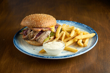 Appetizing American burger with pulled pork, onions, tomatoes, bacon garnished with fries and sauce. Wood background