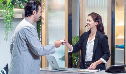 Male guests come in and contact check-in with the front of the counter, there is a hotel reception girl wearing headphones and providing service to deliver the key of the hotel room to the hotel. 