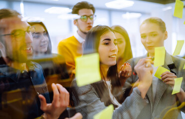 Group of young modern people working on project together. Business colleagues writing on sticky note on glass wall with coworkers standing by in office. Startup Business. Planning, analysis.