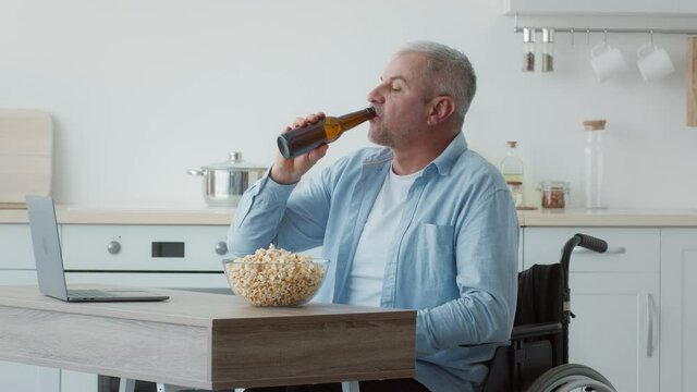 Disabled Man In Wheelchair Using Laptop Drinking Beer At Home