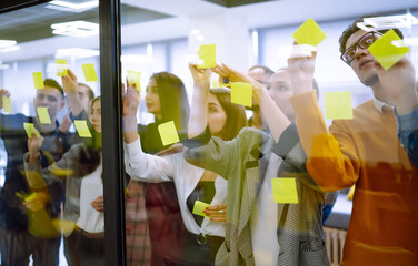 Group of young modern people working on project together. Business colleagues writing on sticky note on glass wall with coworkers standing by in office. Startup Business. Planning, analysis.