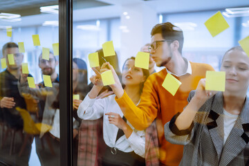 Group of young modern people working on project together. Business colleagues writing on sticky note on glass wall with coworkers standing by in office. Startup Business. Planning, analysis.