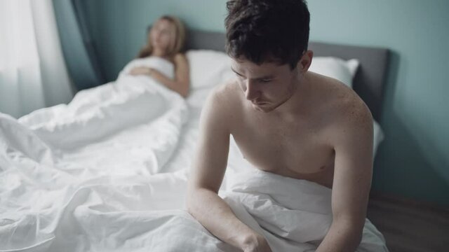 Nervous guy upset sitting on edge of bed because of problems with sex. Young caucasian couple having relationship anxiety. Family conflict. Relationship crisis in couple. Erectile disfunction concept.