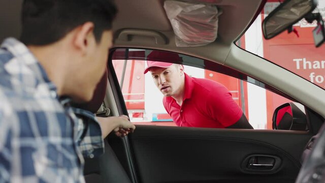 smiling gas station attendant collecting payment through window from young man at driver seat paying with credit card or qr code chatting with him.