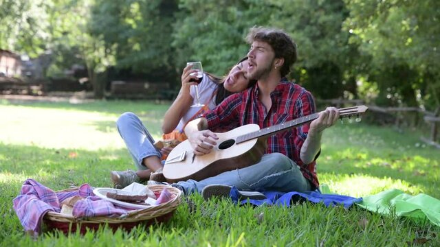 Young caucasian couple, on a sunny day, play the guitar in the park, sing and drink wine.