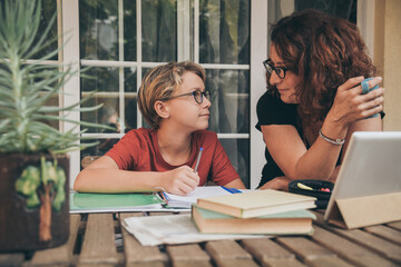 Young student doing homework at home with school books, newspaper and digital pad near his mother. Happy boy looking mum while writing on the copybook. Youth, education and new tech lifestyle concept.