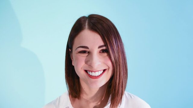 Beautiful caucasian woman with brown hair wearing white shirt starts to smile against blue background. White teeth smile. 4K Resolution video.