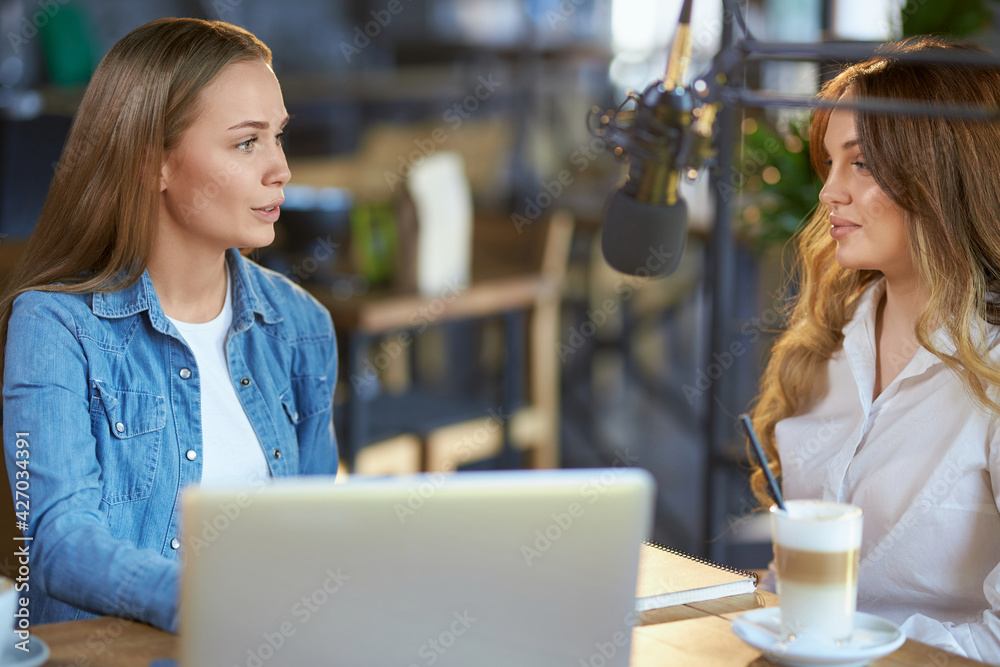 Wall mural Side view of two beautiful young blogger women communicating online with followers or friends in cafe with modern microphone. Concept of process interview with tasty coffee. 