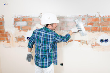 Builder repairman in a protective helmet plastering a brick wall with a putty knife during the...
