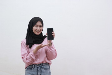 Portrait of a smiling young muslim woman showing blank screen mobile phone isolated over white and minimalist background. Beautiful asian woman with casual dress. Beautiful muslim women.