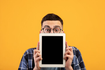 Shocked millennial guy hiding behind tablet computer with empty screen on orange studio background, mockup for design