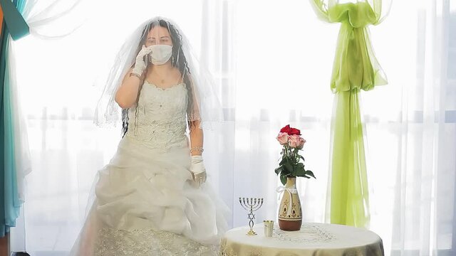 A Jewish bride in a wedding dress and a veil wearing a medical mask in a synagogue hall before the chupah ceremony happily speaks on the phone. Overall plan