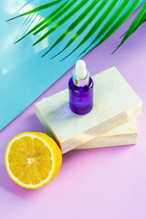 A cosmetic jar with an eyedropper stands on two granite slabs, like on a catwalk, next to half a yellow lemon, a palm leaf and the glare of the sun in the background. Cosmetics for summer