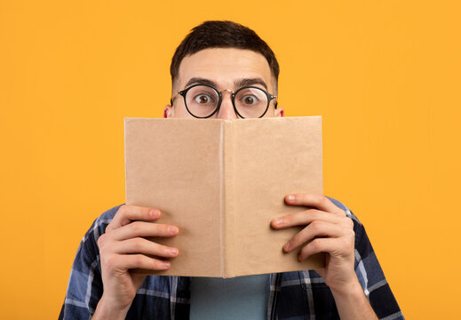 Shocked young guy in glasses hiding behind book, having problem studying for college exams on orange background