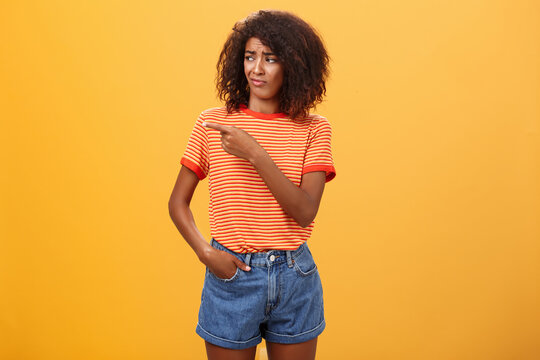 Girl looking at doubtful unimpressive perfomance of model. Portrait of displeased confused good-looking dark-skinned female with afro hairstyle looking and pointing left with scorn and indifference