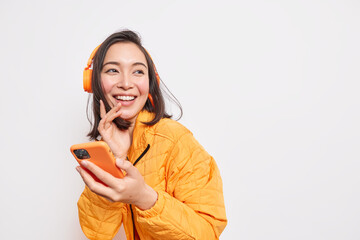 Happy positive Asian female enjoys music uses wireless headphones holds smartphones turns away...