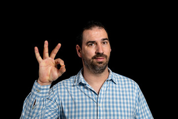 Bearded man in a white and blue plaid shirt doing the OK symbol isolated on black studio background