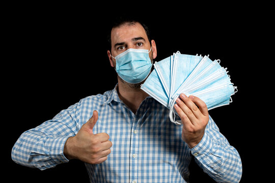 Handsome Bearded Man Wearing A Plaid Shirt And Surgical Mask To Protect Himself From Coronavirus By Lifting His Thumb Up With One Hand And Holding Several Surgical Masks With The Other Hand