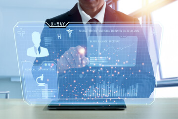 An Asian man wearing a suit points his finger at a 3D monitor that displays blood pressure checkups. Heart rate, blood components Display from the app in the tablet. Blurred building window backdrop
