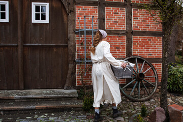 Eine Frau geht mit dem Wäschekorb vom deutschen Fachwerkhaus. Sie hat ein Kopftuch und Bauer Bekleidung an. Ein alter Rad steht vor der Wand.  - Powered by Adobe