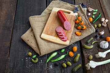 Salami sausage on wooden cutting board with cherry tomatoes, chilli pepper, basilic, garlic, olives, cucumber and spices on vintage wooden background. Copy space.