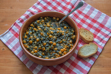 clay pot with chickpeas with spinach with spoon and two pieces of bread on a white and red checkered cloth and wooden background.