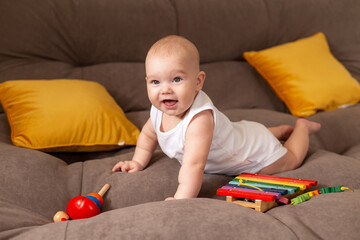 Obraz na płótnie Canvas Cute toddler in white bodysuit lies at home on grey sofa with yellow pillows playing with wooden developing toy