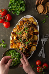 Woman decorates with green onions a dish with stewed wild goat and baked potatoes, Mache salad, bread and tomatoes on a dark background, top view
