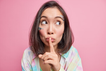 Fototapeta premium Shh dont tell anyone my secret. Mysterious brunette Asian woman presses index finger to lips makes hush sign asks stay quiet shows tabboo gesture concentrated aside poses against rosy studio wall