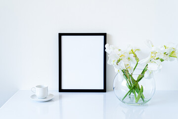 Blank black frame mockup. Fresh flowers of white irises in glass vase in the shape of a sphere on a white table and coffee cup. White background, minimal and elegant space