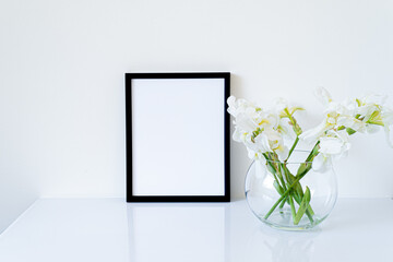 Blank black frame mockup. Fresh white irises flowers in sphere shaped glass vase on white table. White background, minimal and elegant space
