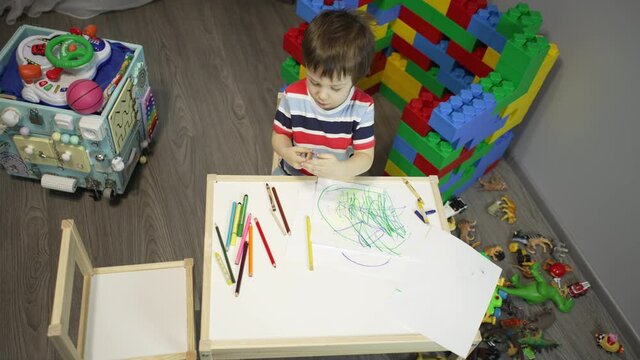 The boy draws pictures at the children's table in the room. The boy is engaged in education and creativity drawing a picture. Beautiful child at home studying.