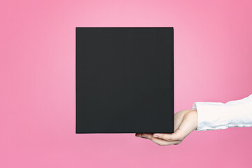 A square black box with an copy empty space for text and design in the hand of a young woman in a white shirt on a pink background. Board, blank, template, mockup, layout for a slogan or inscription.