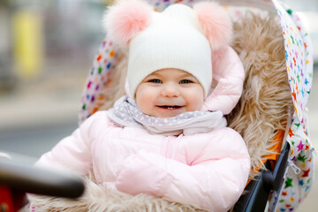 Cute little beautiful baby girl sitting in the pram or stroller on cold autumn, winter or spring day. Happy smiling child in warm clothes, fashion stylish baby coat and hat. Snow falling down