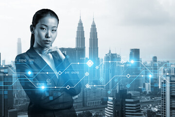 Attractive young Asian businesswoman pondering on technology at business process to achieve tremendous growth in commerce. Tech hologram icons over Kuala Lumpur background