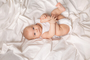 Cute toddler in white bodysuit lies in bed with white bedding