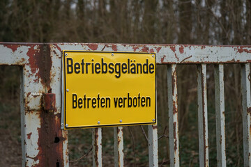 "Betriebsgelände - Betreten verboten": Gelbes Hinweisschild vor einem Privatgrundstück an einem rostigen Zauntor 