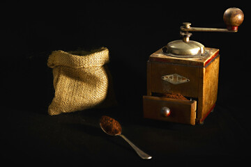Photo composition with an antique coffee grinder, a hemp bag and a silver spoon with ground coffee on a black background