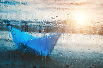 Blurry background with sunlight on water of blue paper boat washed up on beach after rain behind wet glass in raindrops.
