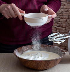 Hands hold a sieve, sifting flour into a bowl of dough. Next to it is a mixer for mixing. Selective focus.