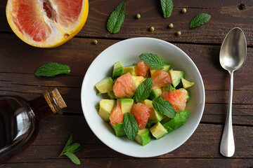 Salad with avocado, grapefruit and mint, a spoon, a grapefruit and a bottle with vegatable oil on a dark wooden table, top view