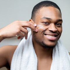 Obraz premium Man facial care. Cosmetic product. Metrosexual lifestyle. Cheerful shirtless African guy with white towel applying face cream with fingers smiling isolated on light background.