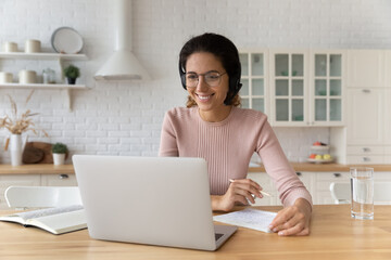 Pleasant elearning. Hispanic female student in earphones study from home look on pc screen listen to tutor teacher prepare to take notes. Smiling young woman enjoy watching webinar online using laptop - obrazy, fototapety, plakaty