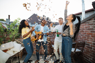 Group of four multiracial people in casual clothes drinking alcohol, playing guitar, singing and throwing up confetti. Concept of party time and enjoyment.