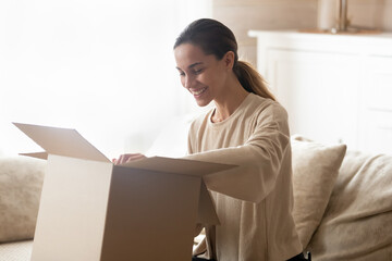Smiling millennial Hispanic woman unbox cardboard package shopping online from home. Happy young female buyer feel excited with good delivery service unpack open box with internet order.