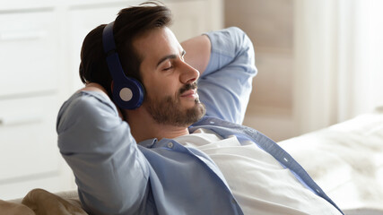 Close up of calm young Caucasian man in headphones relax on sofa at home listen to music take nap. Millennial male in earphones rest on couch breathe fresh air, enjoy good quality sound on device.