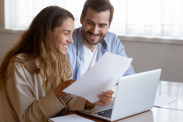 Smiling young multiethnic man and woman sit at desk at home work on computer read good news in paperwork letter. Happy couple use laptop pay bills excited with pleasant message in correspondence.