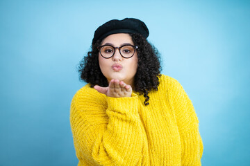Young woman wearing french look with beret and yellow casual sweater over isolated blue background looking at the camera blowing a kiss with hand on air being lovely and sexy. Love expression.